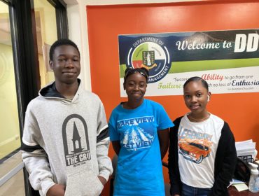 Three teens stand smiling in front of the DDM reception desk